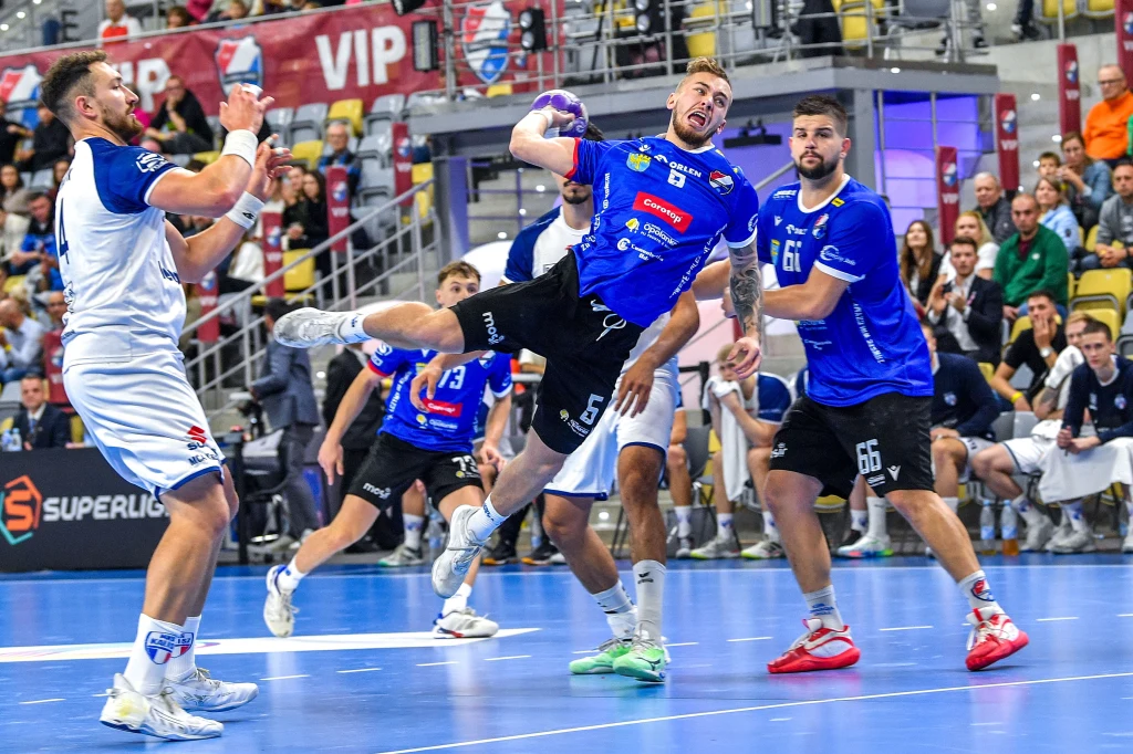 Orlen Superliga Drużyna w niebieskich strojach walczy o przewagę nad zespołem w białych koszulkach podczas dynamicznego meczu piłki ręcznej, zawodnik w wyskoku oddaje rzut na bramkę w obecności licznej publiczności.
