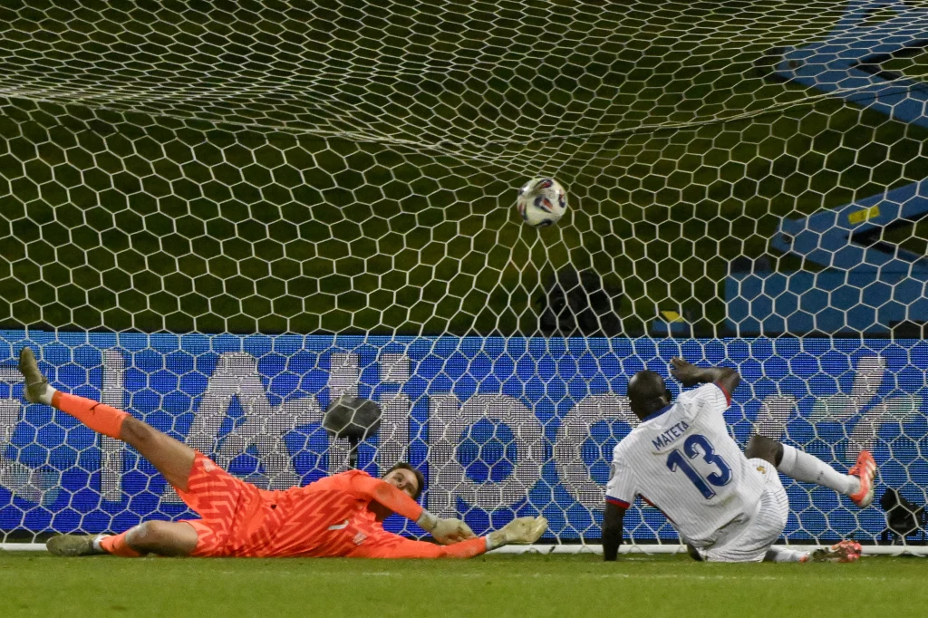 Kiedy do siatki trafił Jean-Philippe Mateta (nr 13), tylko przez moment wydawało się, że Francuzi sięgną po komplet punktów Bramkarz w pomarańczowym stroju rzuca się w lewo próbując obronić piłkę lecącą do bramki podczas rzutu karnego, zawodnik w białym stroju z numerem 13 oddaje strzał, dynamiczna scena na tle siatki bramkowej.