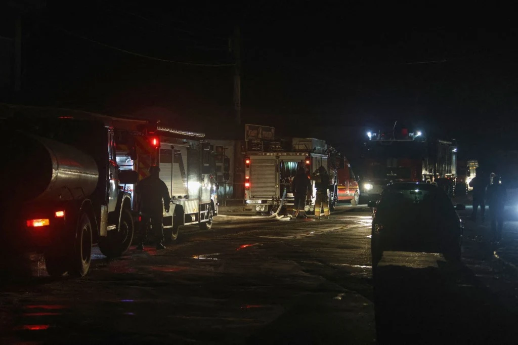 Wozy strażackie ustawione na ciemnej ulicy, kilku strażaków pracuje przy pojazdach, scena oświetlona światłami pojazdów i reflektorami, ogólna atmosfera nocnej akcji ratunkowej.
