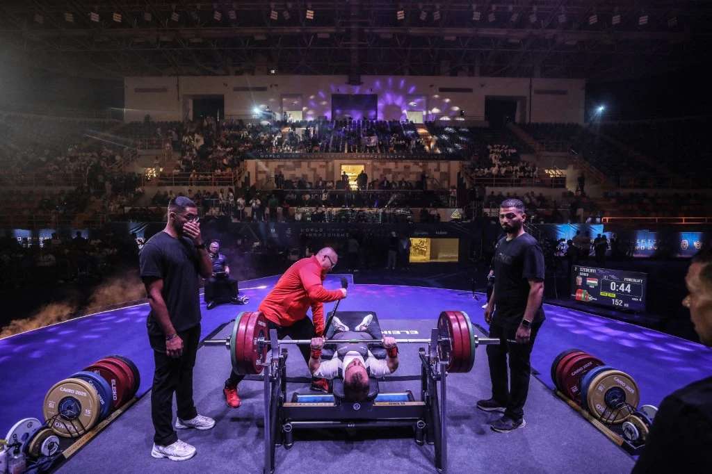Skandal z udziałem Rosji. Sceny na mistrzostwach świata. To naprawdę się wydarzyło Zawody wyciskania sztangi na ławce w hali sportowej, zawodnik podnoszący obciążoną sztangę, otoczony przez sędziów i asystentów, publiczność na trybunach obserwuje przebieg wydarzenia