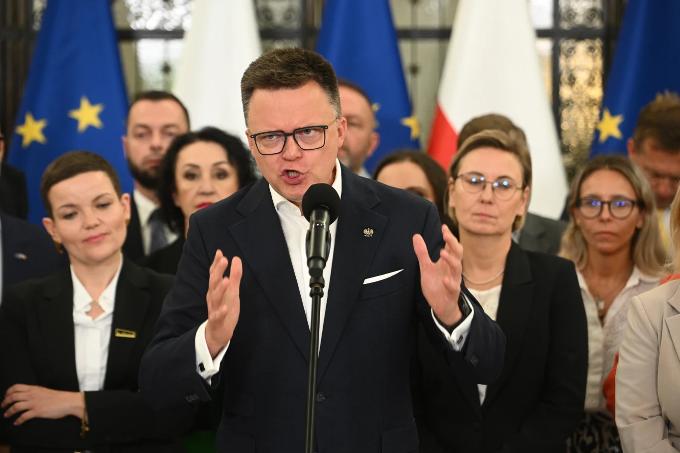 Polska 2050 rozpadnie się bez Szymona Hołowni? Wyniki nowego sondażu Mężczyzna w garniturze przemawia do mikrofonu, gestykulując rękami, w tle znajduje się grupa ludzi oraz flagi Unii Europejskiej i Polski.