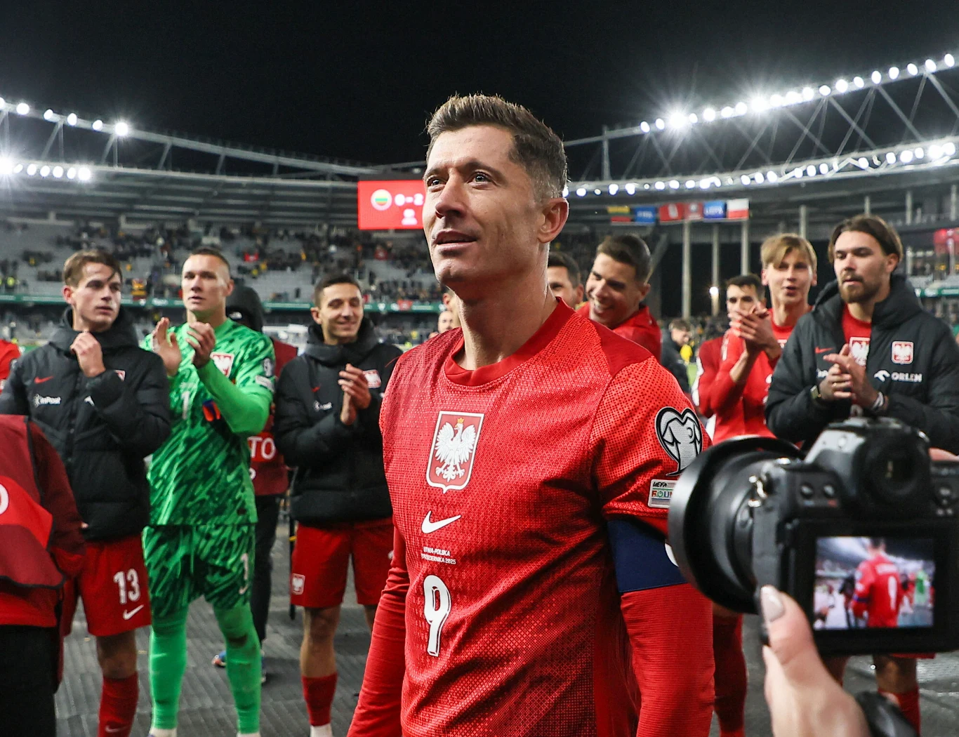 Piłkarz ubrany w czerwoną koszulkę reprezentacji Polski z numerem 9 stoi na stadionie, otoczony przez kolegów z drużyny oraz fotografów, wokół widać trybuny i światła stadionu, na twarzach zawodników widać radość i dumę po zakończonym meczu.