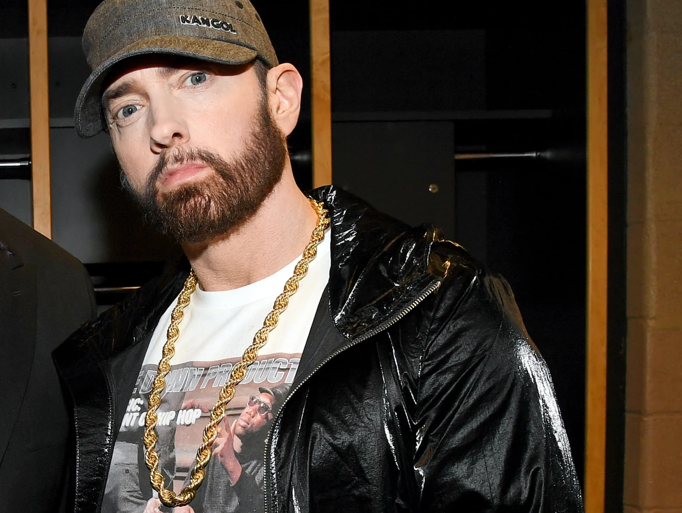 Eminem po raz kolejny zostanie dziadkiem! Mężczyzna w szarej czapce z daszkiem i ciemnej kurtce, noszący biały t-shirt z nadrukiem oraz złoty łańcuch na szyi, stoi na tle wnętrza budynku.
