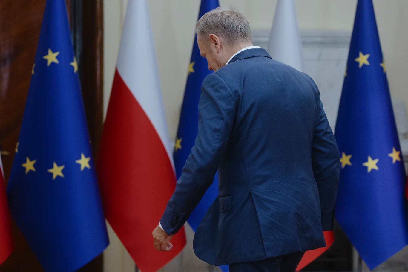 Co zostało z wygranej Koalicji 15 października? Mężczyzna w garniturze odwrócony plecami, przechodzący obok ustawionych naprzemiennie flag Polski oraz Unii Europejskiej w oficjalnym wnętrzu.