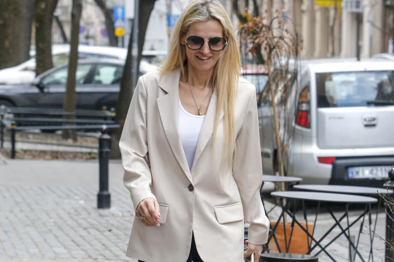 Uśmiechnięta kobieta w jasnej marynarce i białym topie idzie chodnikiem miejskiej ulicy, nosi okulary przeciwsłoneczne i ma rozpuszczone blond włosy. W tle widoczne są zaparkowane auta oraz elementy miejskiego krajobrazu.