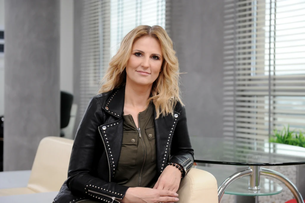 Dziś Dominika Tajner już tak nie wygląda Kobieta o blond włosach siedząca na jasnej sofie w nowoczesnym biurze; ubrana w czarną skórzaną kurtkę z ozdobnymi ćwiekami i oliwkową bluzkę, za nią widać duże okna z żaluzjami oraz szklany stolik.