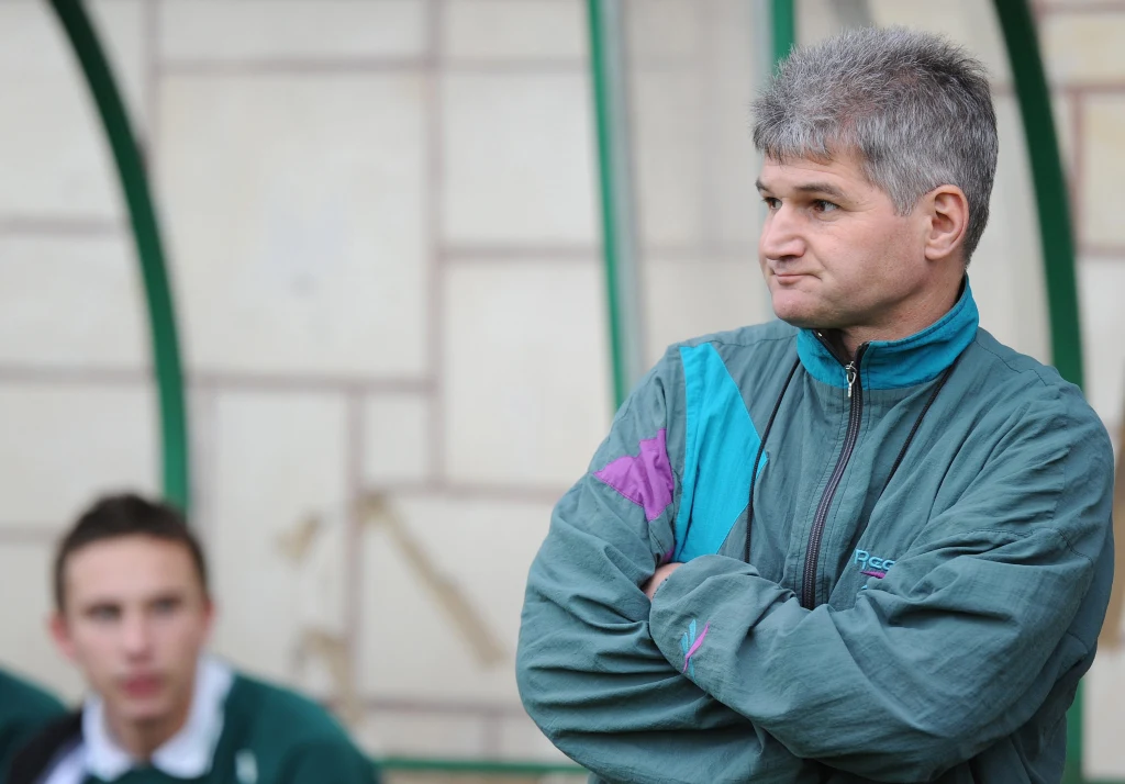 Krzysztof Urban Mężczyzna w średnim wieku w kurtce sportowej stoi z założonymi rękami na tle murku, za nim w tle widoczny młody mężczyzna w zielonej bluzie, obraz sugeruje atmosferę oczekiwania lub skupienia.