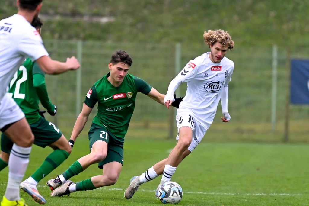 Dwóch piłkarzy rywalizuje o piłkę na murawie podczas meczu piłki nożnej, zawodnik w białym stroju prowadzi piłkę, a zawodnik w zielonym próbuje go powstrzymać.