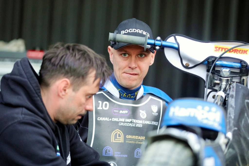 Zafrasowany Jarosław Hampel. Obok jego wieloletni mechanik Michał Przybylski. Dwóch mężczyzn w otoczeniu motocykla żużlowego, jeden z nich ubrany w kevlar sportowy i czapkę z logotypem, drugi pochylony nad motocyklem wykonuje czynność serwisową.