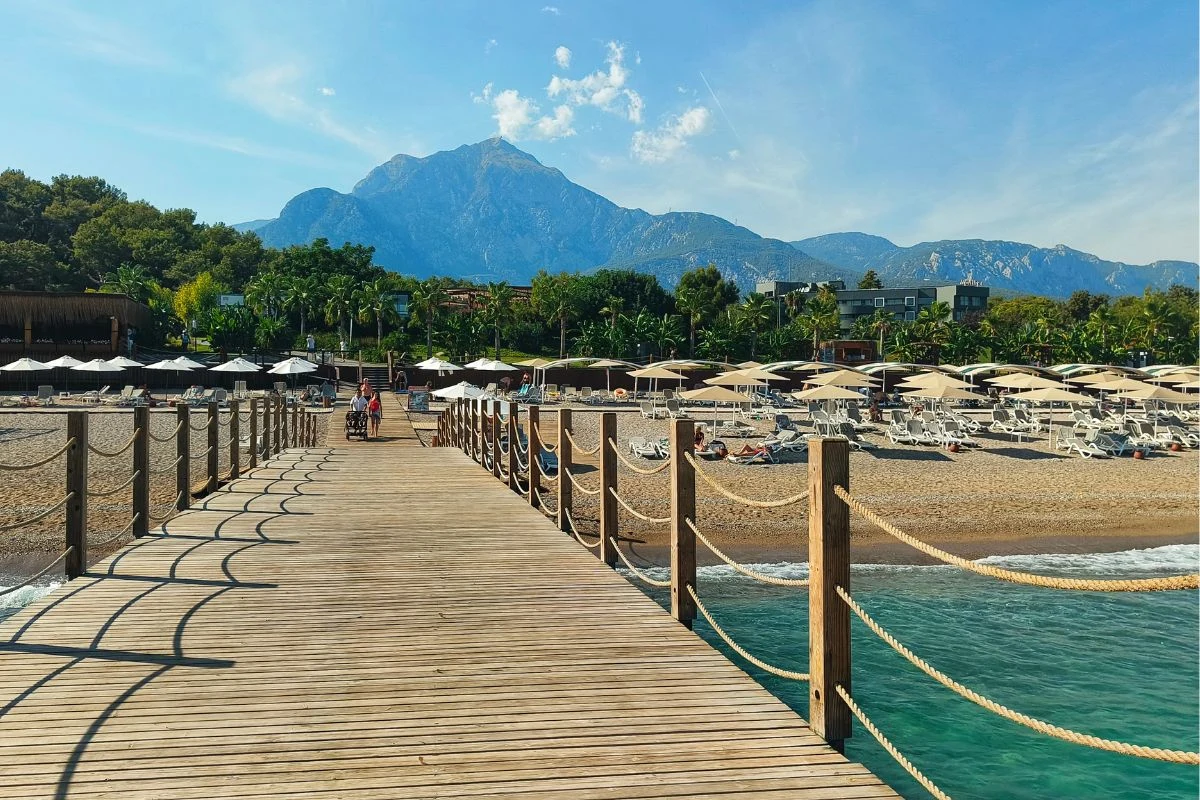 Drewniane molo prowadzące wprost na piaszczystą plażę z licznymi leżakami i parasolami, w tle góry porośnięte zielenią oraz niebieskie niebo z lekkimi chmurami.