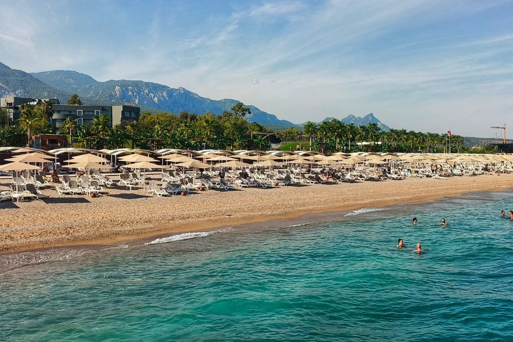 Plaża w miejscowości Tekirova Plaża żwirowa z licznymi leżakami i parasolami, otoczona przez soczystą zieleń palm i zabudowania hotelowe, w tle pasmo górskie, a w wodzie kąpią się ludzie.