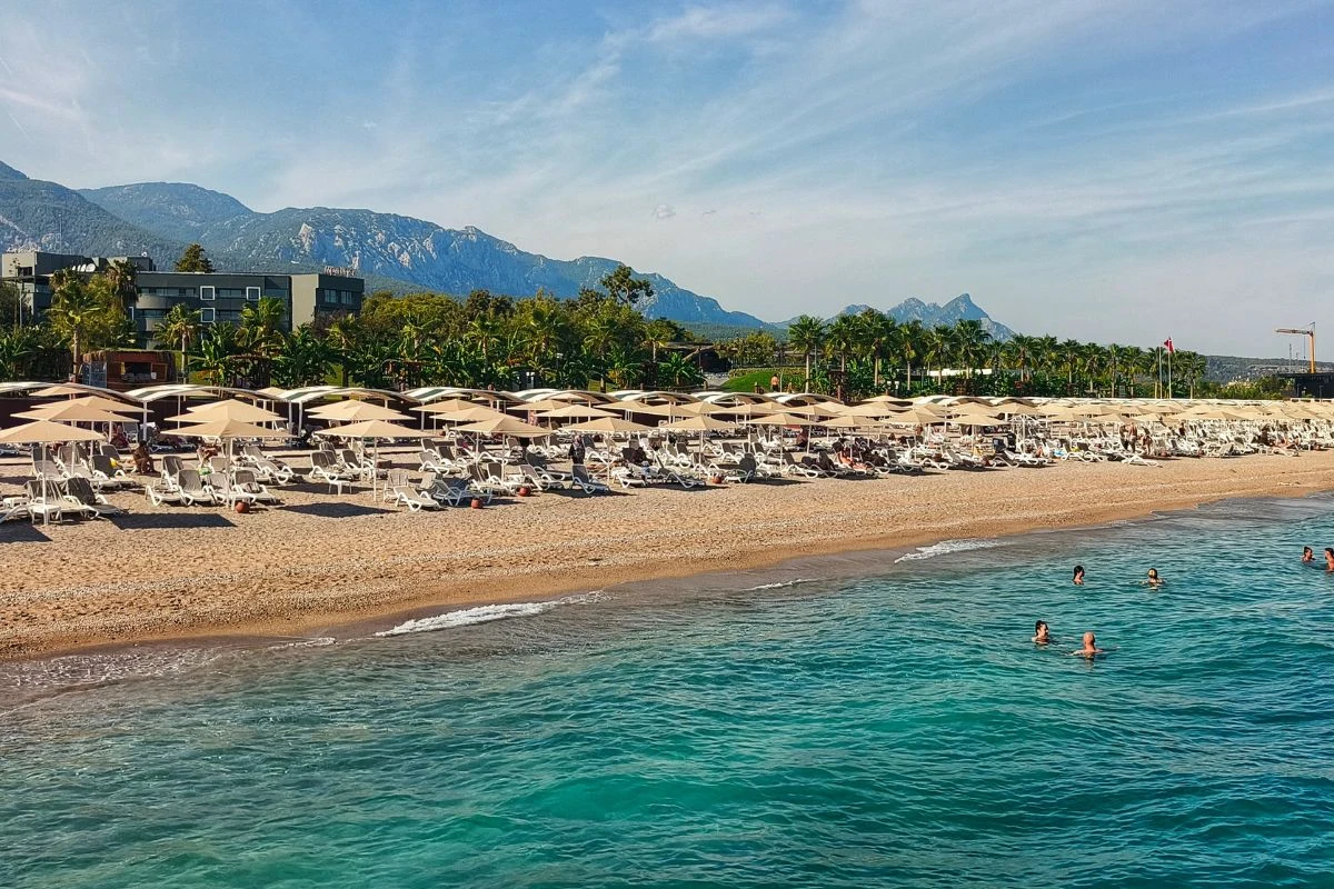 Plaża żwirowa z licznymi leżakami i parasolami, otoczona przez soczystą zieleń palm i zabudowania hotelowe, w tle pasmo górskie, a w wodzie kąpią się ludzie.
