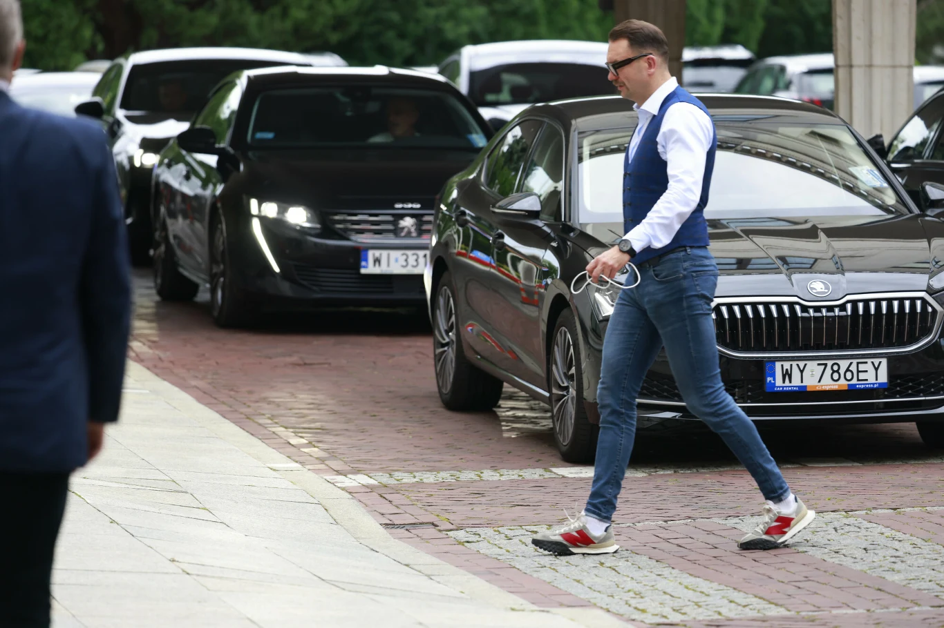 Elegancko ubrany mężczyzna w niebieskiej kamizelce, białej koszuli i dżinsach idzie chodnikiem obok luksusowych czarnych samochodów zaparkowanych przy ulicy.