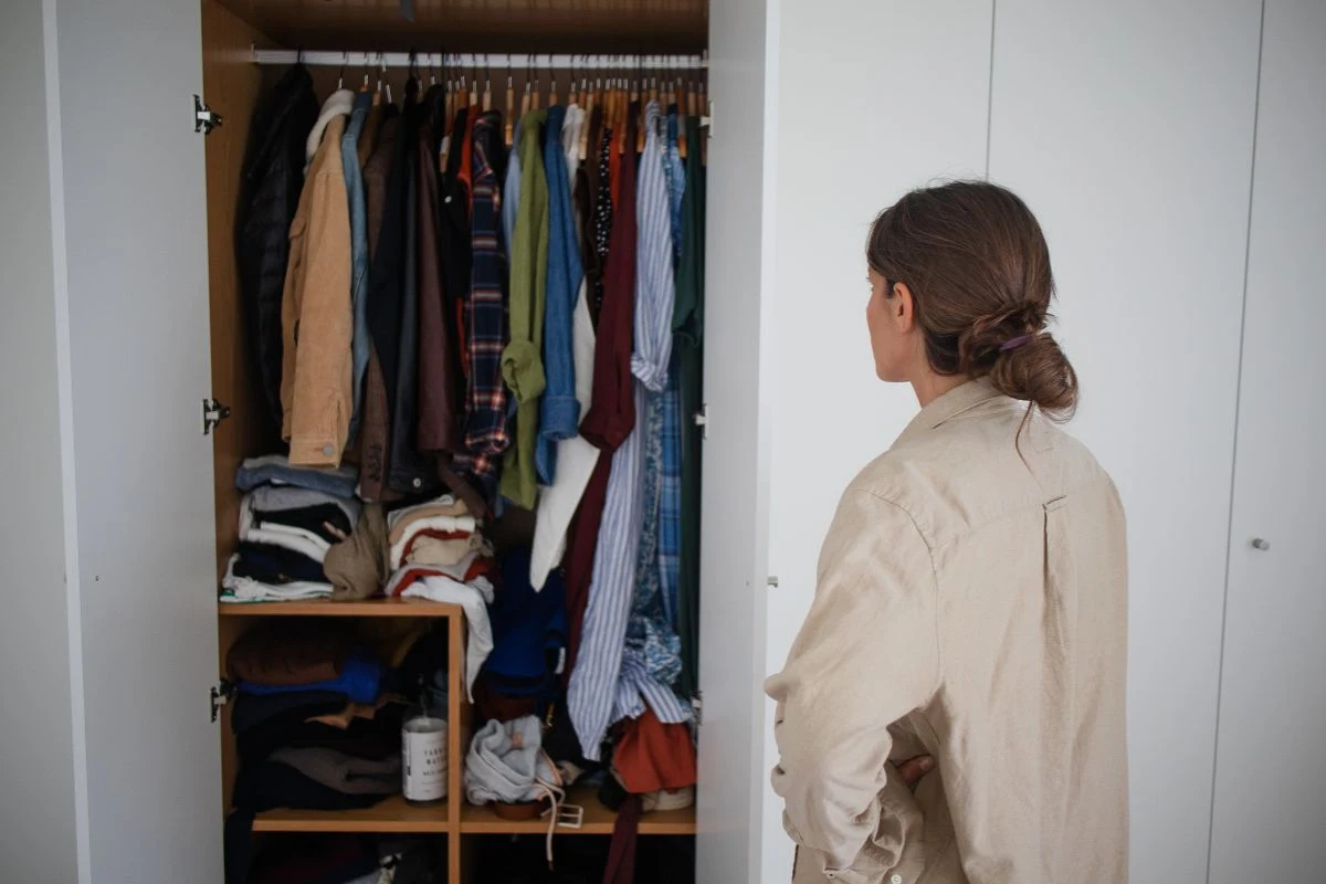 Osoba stojąca tyłem przed otwartą szafą pełną ubrań na wieszakach oraz półkach, ubrania ułożone w różnych pozycjach, światło dzienne oświetla główną scenę.