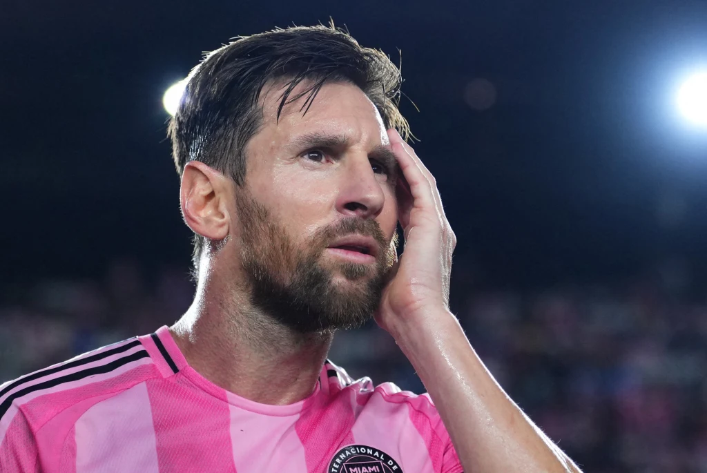 Lionel Messi Piłkarz w różowej koszulce z logo klubu na piersi na tle stadionowego oświetlenia trzyma dłoń na twarzy, wydaje się zamyślony lub zaniepokojony, w tle rozmyte światła i niewyraźne sylwetki.