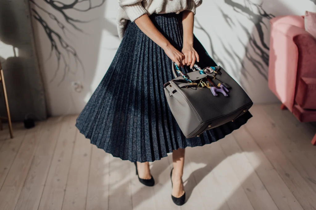 Plisowana spódnica doda szyku i elegancji Kobieca sylwetka ubrana w plisowaną granatową spódnicę i jasny sweter, trzymająca elegancką torebkę z ozdobami, stojąca w jasnym, stylowym wnętrzu na tle rozmytych cieni.