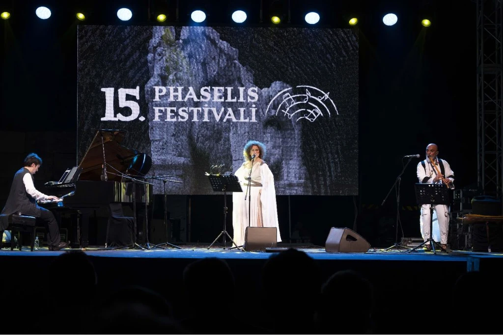 Muzyczna uczta zorganizowana w starożytnym mieście smakuje wyśmienicie Trójka artystów w eleganckich strojach wykonuje utwory muzyczne na scenie podczas festiwalu, w tle widoczny jest duży ekran z napisem „15. Phaselis Festivali” i grafiką przedstawiającą ruinę.