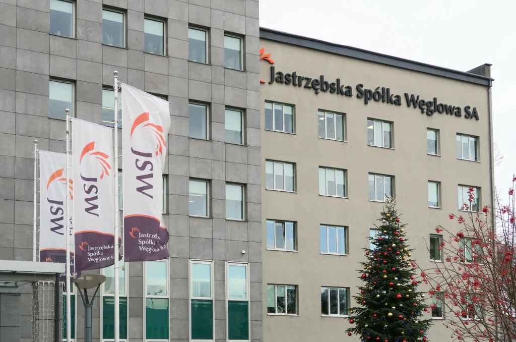 Zarząd JSW przygotowuje trudną restrukturyzację. Zredukowane mają być koszty pracy. Zdj. ilustracyjne Budynek firmy Jastrzębska Spółka Węglowa SA z widocznymi logotypami na fasadzie oraz na flagach przed wejściem, obok dekoracyjna choinka świąteczna z ozdobami.