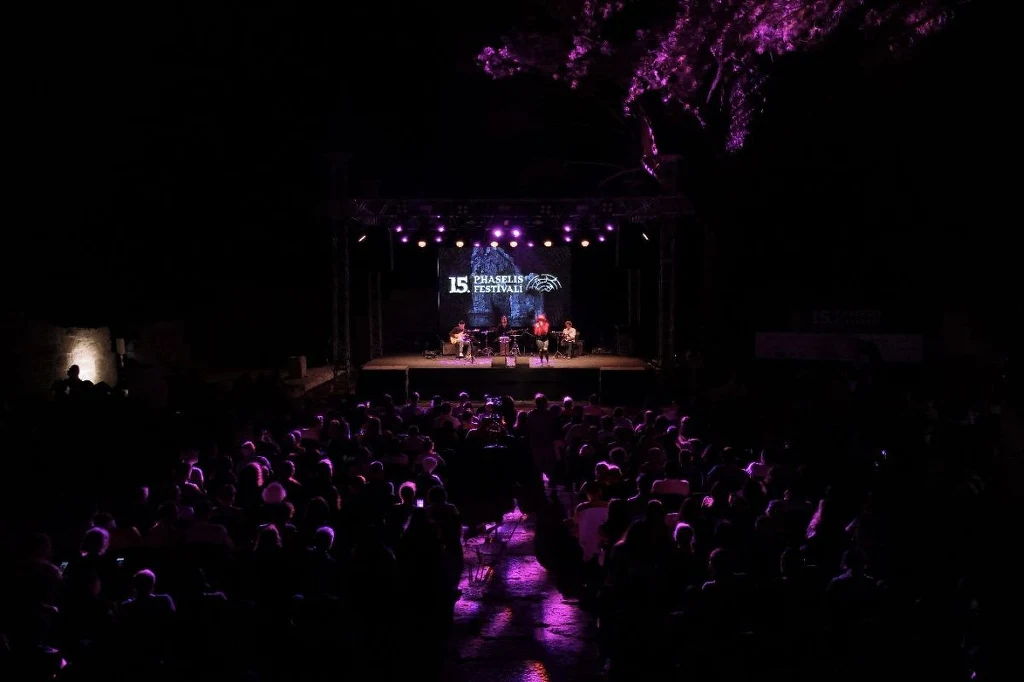 15. Phaselis Festival przyciągnął wielu melomanów Widownia siedząca przed sceną na świeżym powietrzu, oświetloną fioletowymi światłami, na scenie kilka osób bierze udział w panelu festiwalowym, nad nimi widoczny jest napis wskazujący na festiwal literacki.
