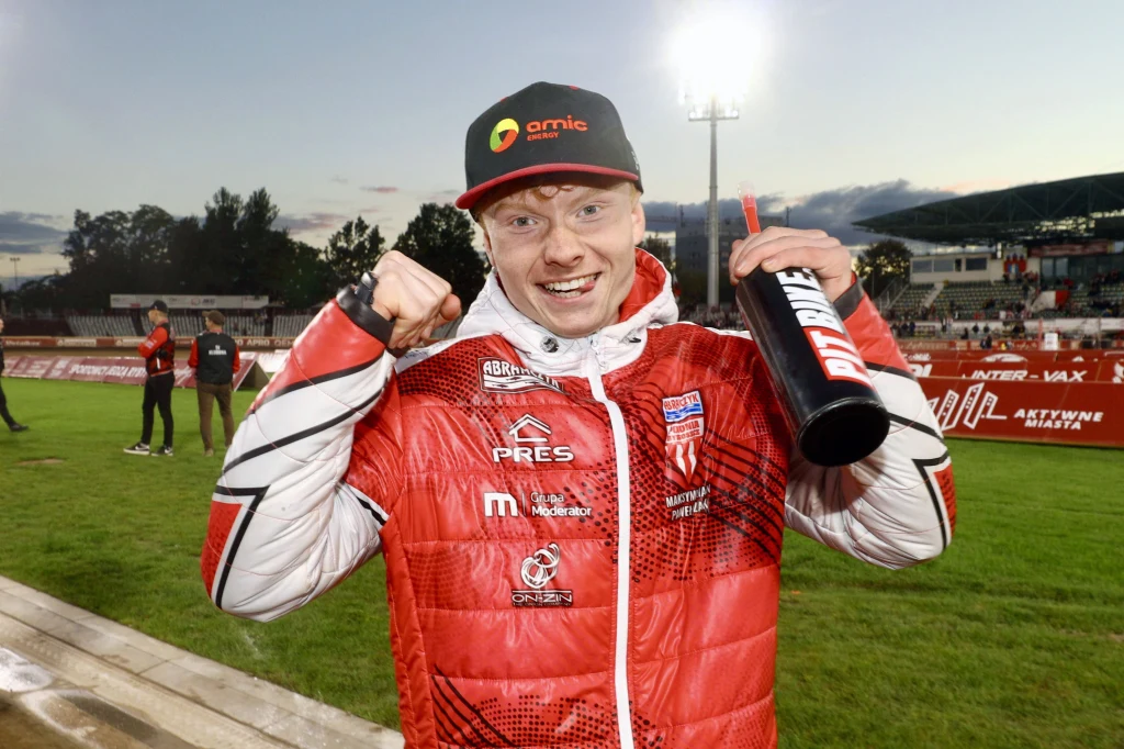 Młody mężczyzna w czerwonej kurtce sportowej i czapce z napisem, uśmiechnięty, trzyma w ręce bidon i wykonuje gest zwycięstwa zaciśniętą pięścią, w tle stadion sportowy z publicznością oraz innymi uczestnikami.