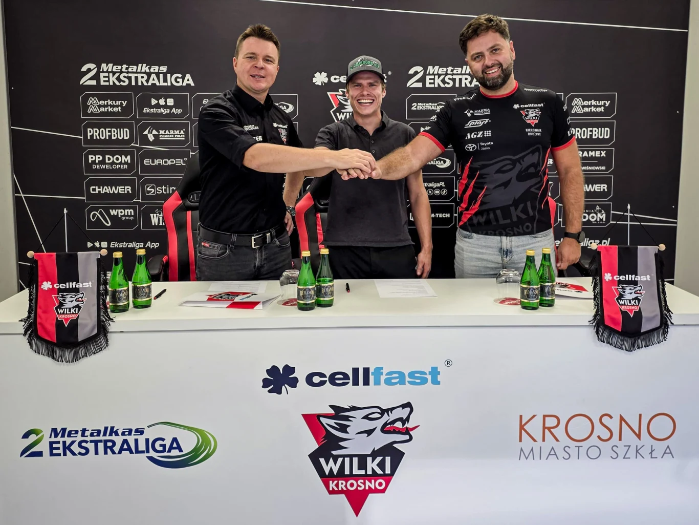 Trzech mężczyzn zawierających umowę przy stole, na którym znajdują się butelki z wodą, apasze klubowe i materiały promocyjne Wilki Krosno oraz sponsorów, w tle ścianka z logotypami partnerów i sponsorów.
