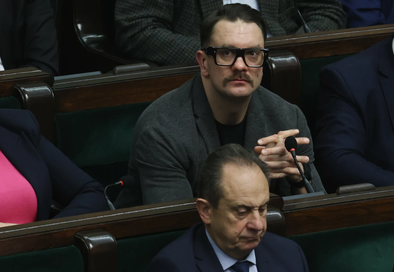 Łukasz Mejza Mężczyzna w okularach i z wąsem siedzi na sali sejmowej wśród innych osób, w tle widoczne drewniane ławki oraz mikrofony.
