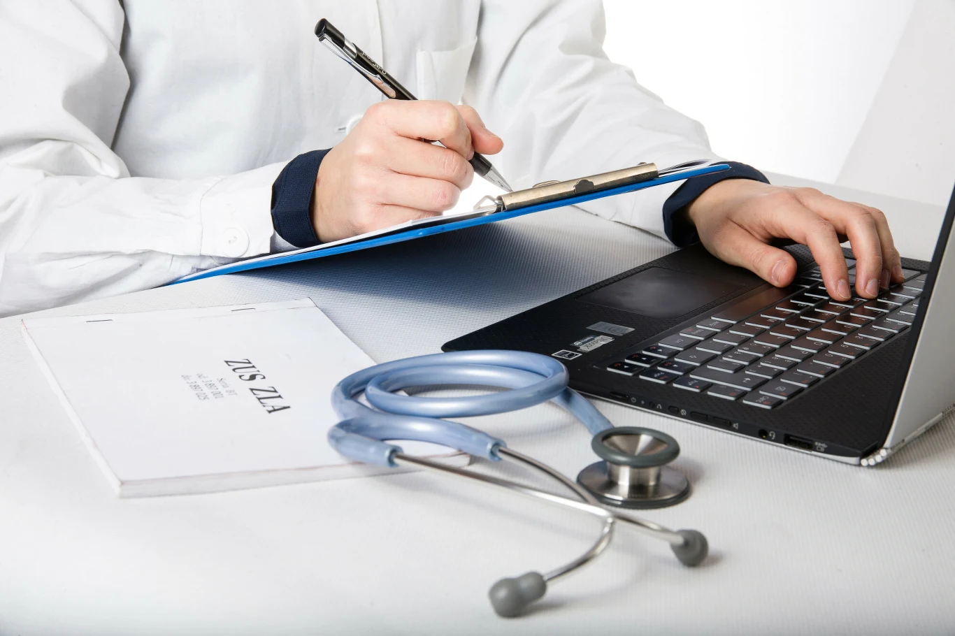 W kwietniu wchodzą zmiany w przepisach dotyczących L4. Lekarz w białym fartuchu zapisuje notatki na niebieskim klipbordzie, przed nim znajduje się otwarty laptop, stetoskop i dokumentacja medyczna.