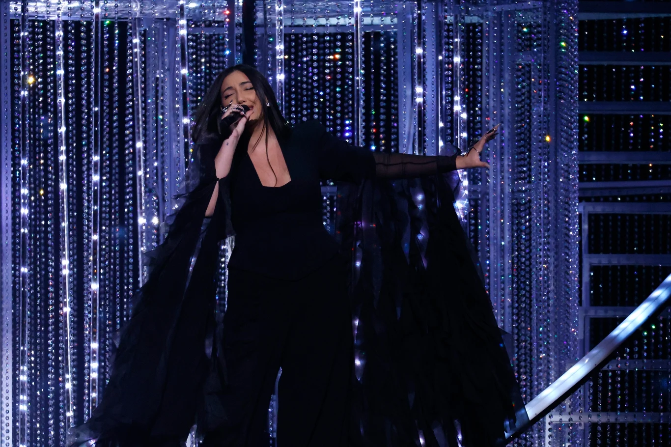 Juwal Rafael (Izrael) podczas Konkursu Piosenki Eurowizji 2025 Kobieta ubrana na czarno śpiewa na scenie, w tle połyskujące kurtyny z kryształków i metalowa konstrukcja, dynamiczna poza i ekspresja artystyczna podczas występu.