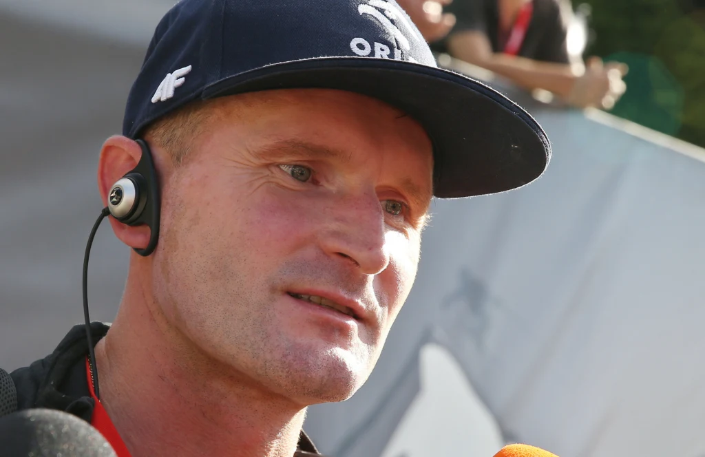 Maciej Maciusiak Mężczyzna w czapce z logo Orlen oraz słuchawką w uchu podczas udzielania wywiadu, w tle widoczne zamazane postacie i fragmenty transparentu.