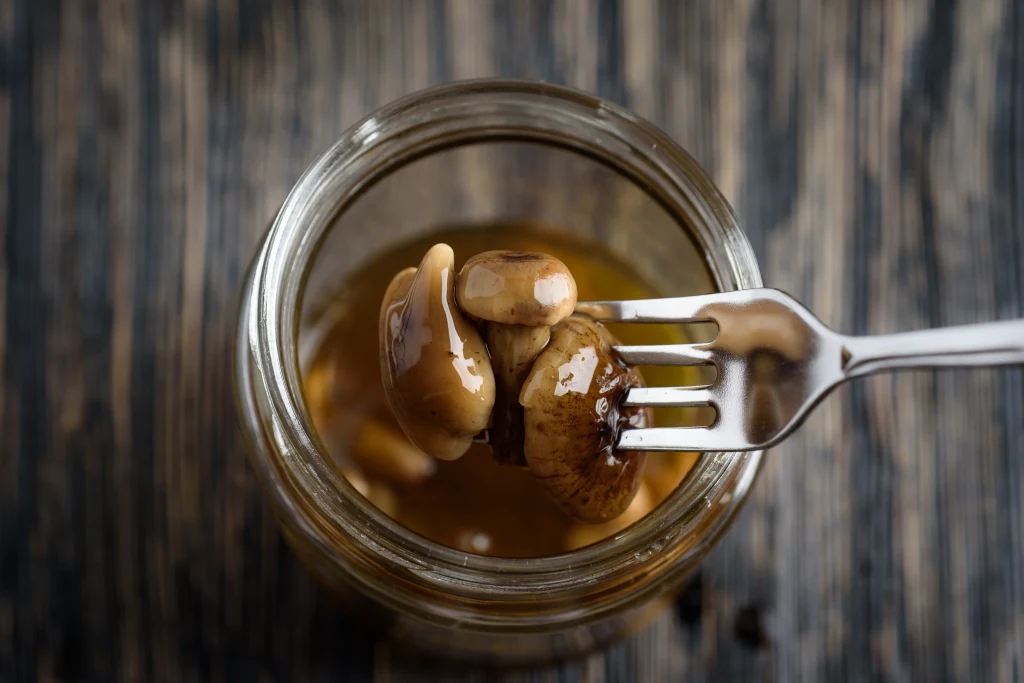 Widelec trzyma marynowane grzyby wyjmowane ze słoika wypełnionego zalewą, na tle drewnianego blatu.