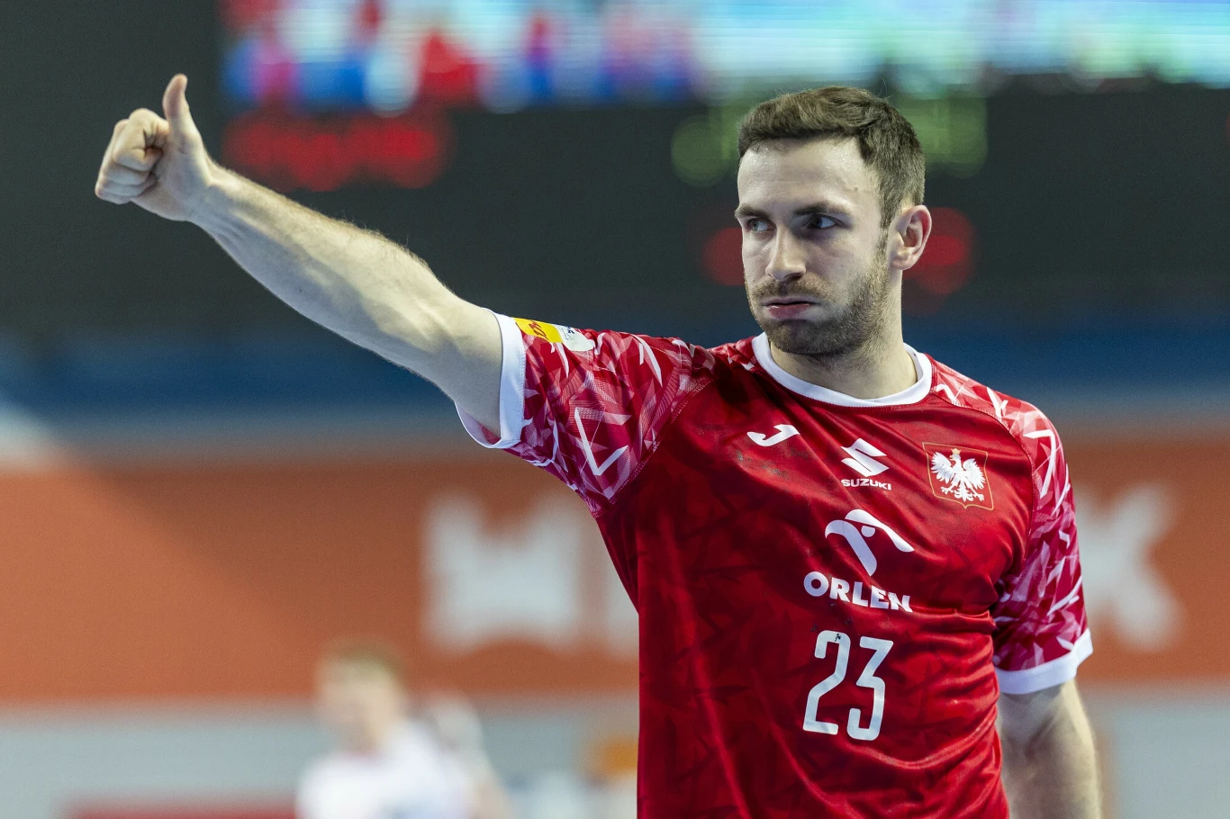 Zawodnik piłki ręcznej w czerwonej koszulce z numerem 23 unosi kciuk do góry podczas meczu, na koszulce widoczne logo sponsora oraz herb kraju.