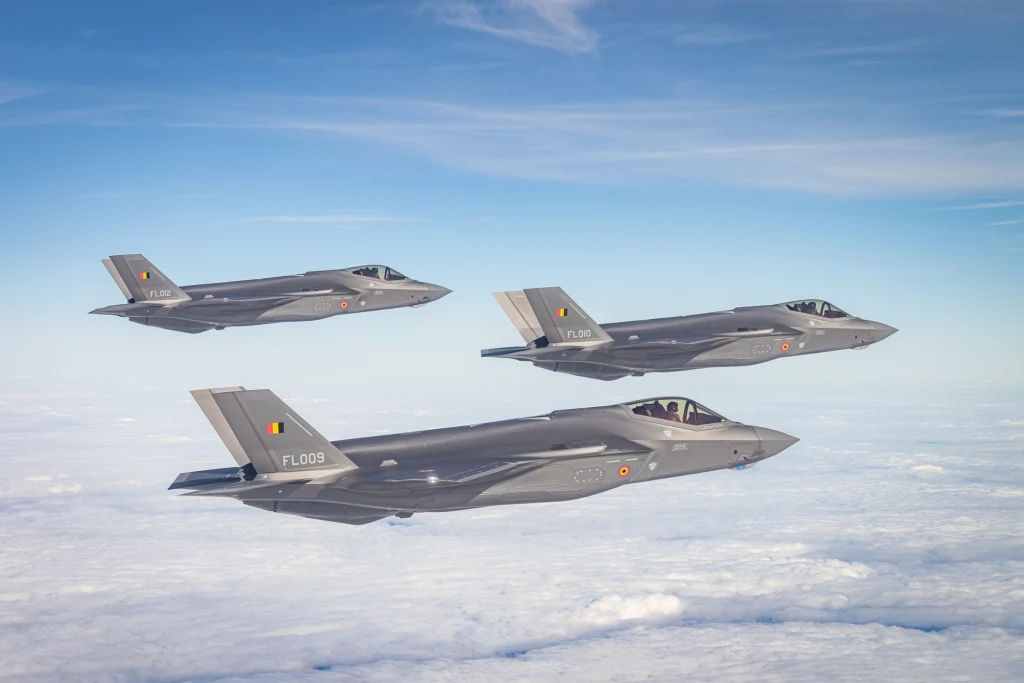 Belgijskie F-35 lecące do kraju. Jak widać, zostały ozdobione kolorowymi symbolami krajowych sił powietrznych Trzy nowoczesne myśliwce F-35 w szyku podczas lotu powyżej poziomu chmur na tle błękitnego nieba, każdy z charakterystycznymi oznaczeniami i flagą Belgii na ogonie.