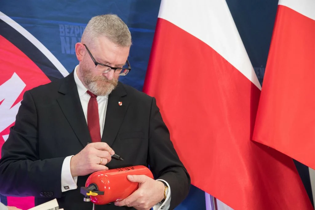 Grzegorz Braun podpisujący gaśnicę Mężczyzna w garniturze podpisuje czerwony przedmiot przypominający sprzęt gaśniczy, w tle widoczne polskie flagi oraz fragment logotypu.
