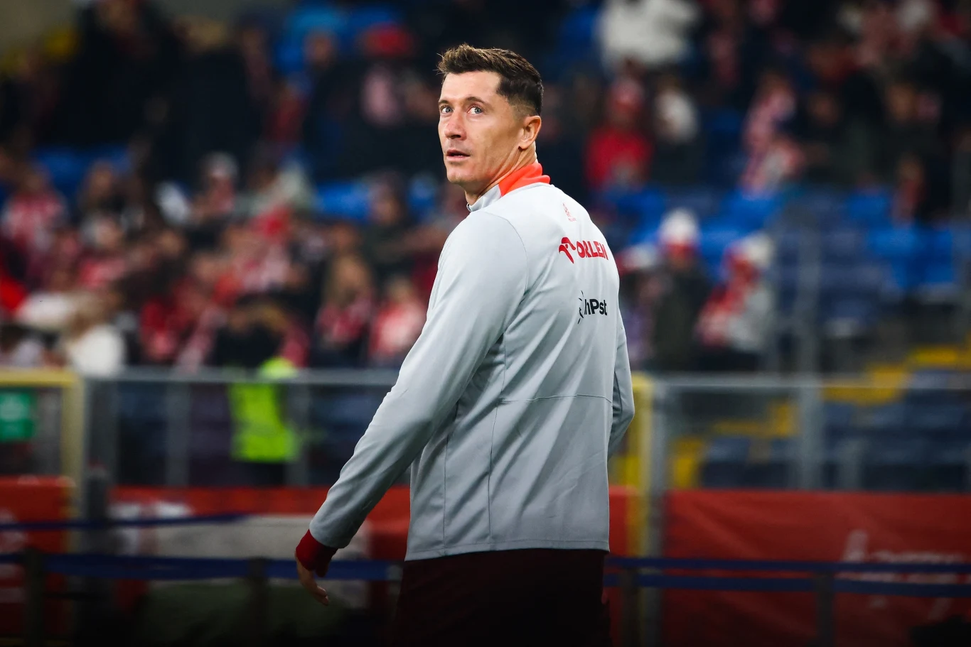 Robert Lewandowski Piłkarz w sportowym stroju z napisem Polska na stadionie, skupiony, z trybunami i kibicami w tle.