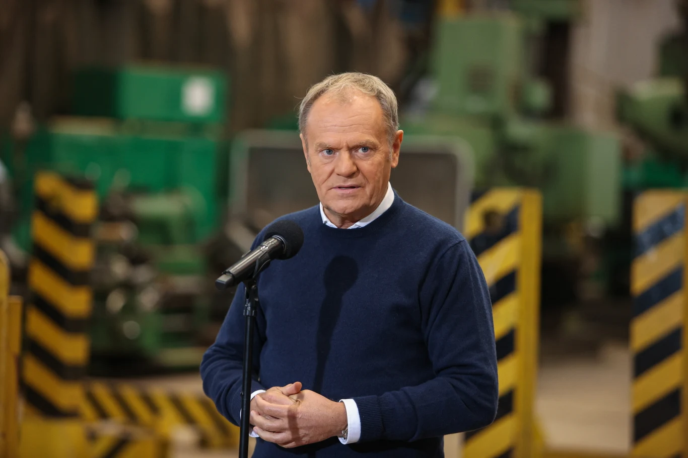 Premier Donald Tusk podczas wtorkowej konferencji prasowej w Raciborzu Mężczyzna w niebieskim swetrze przemawia do mikrofonu na tle przemysłowej hali z maszynami oraz barierami w żółto-czarne pasy.