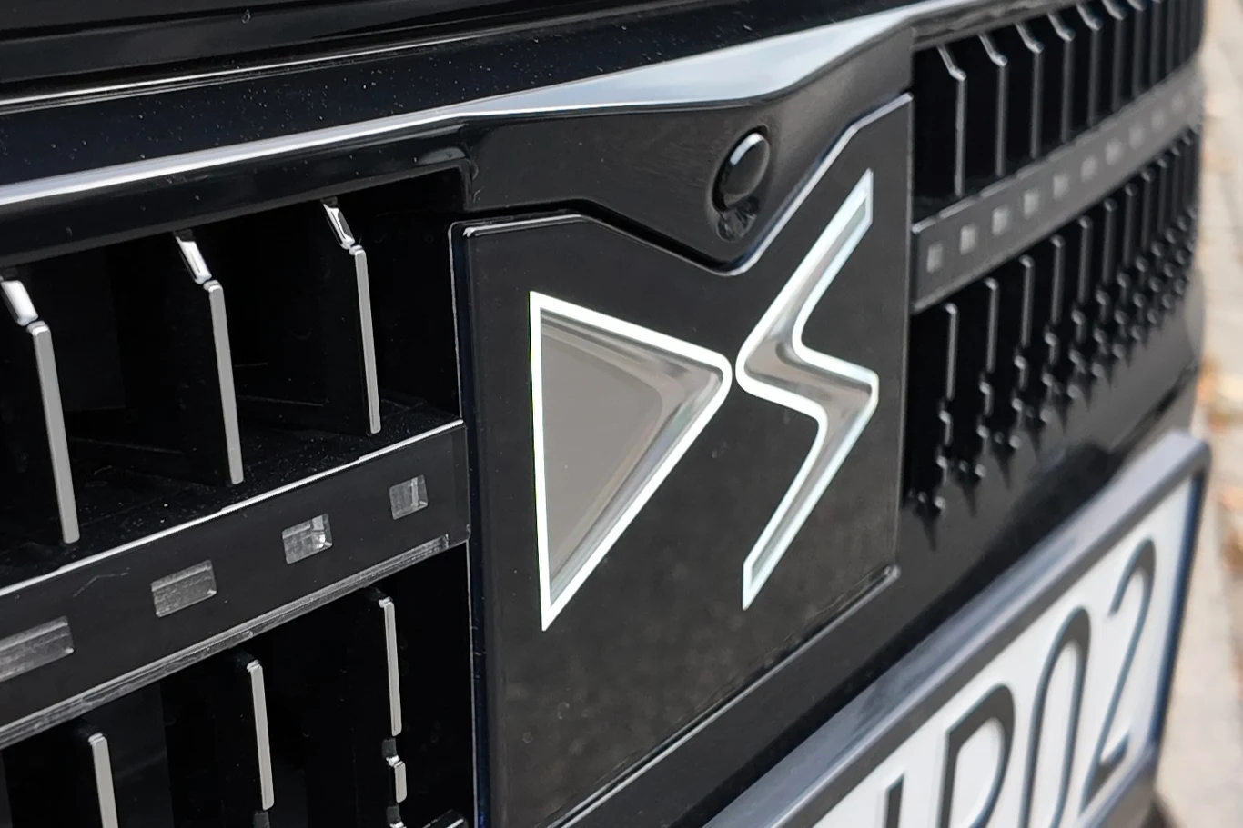 Zbliżenie na przednią część samochodu z widocznym logo marki DS oraz fragmentem tablicy rejestracyjnej. Chromowane elementy kratki i czarne lakierowane powierzchnie podkreślają nowoczesny design pojazdu.