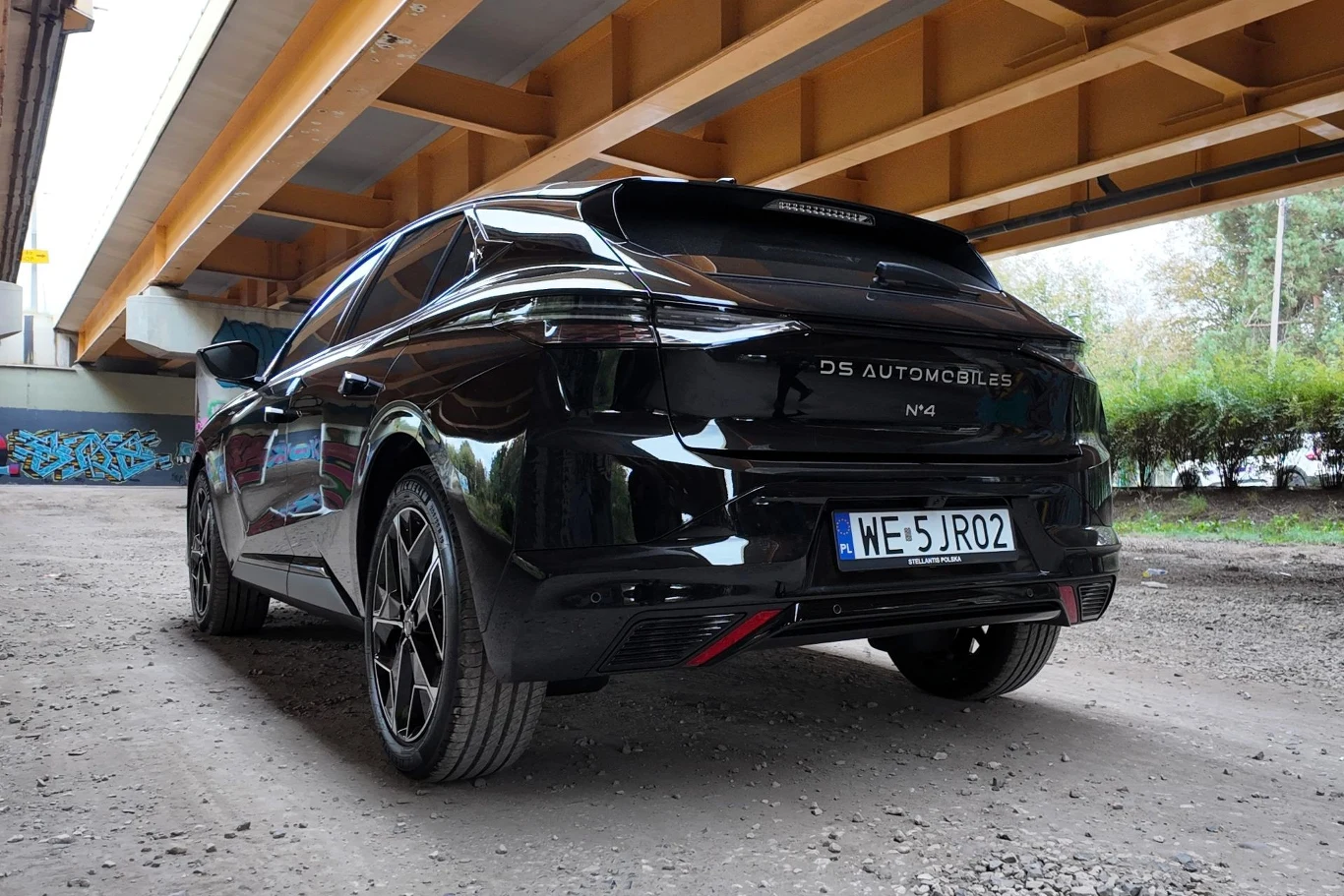 Czarny samochód marki DS Automobiles, model DS4 stoi zaparkowany pod wiaduktem na nieutwardzonym terenie, w tle widoczne są graffiti na betonowych podporach oraz krzewy.