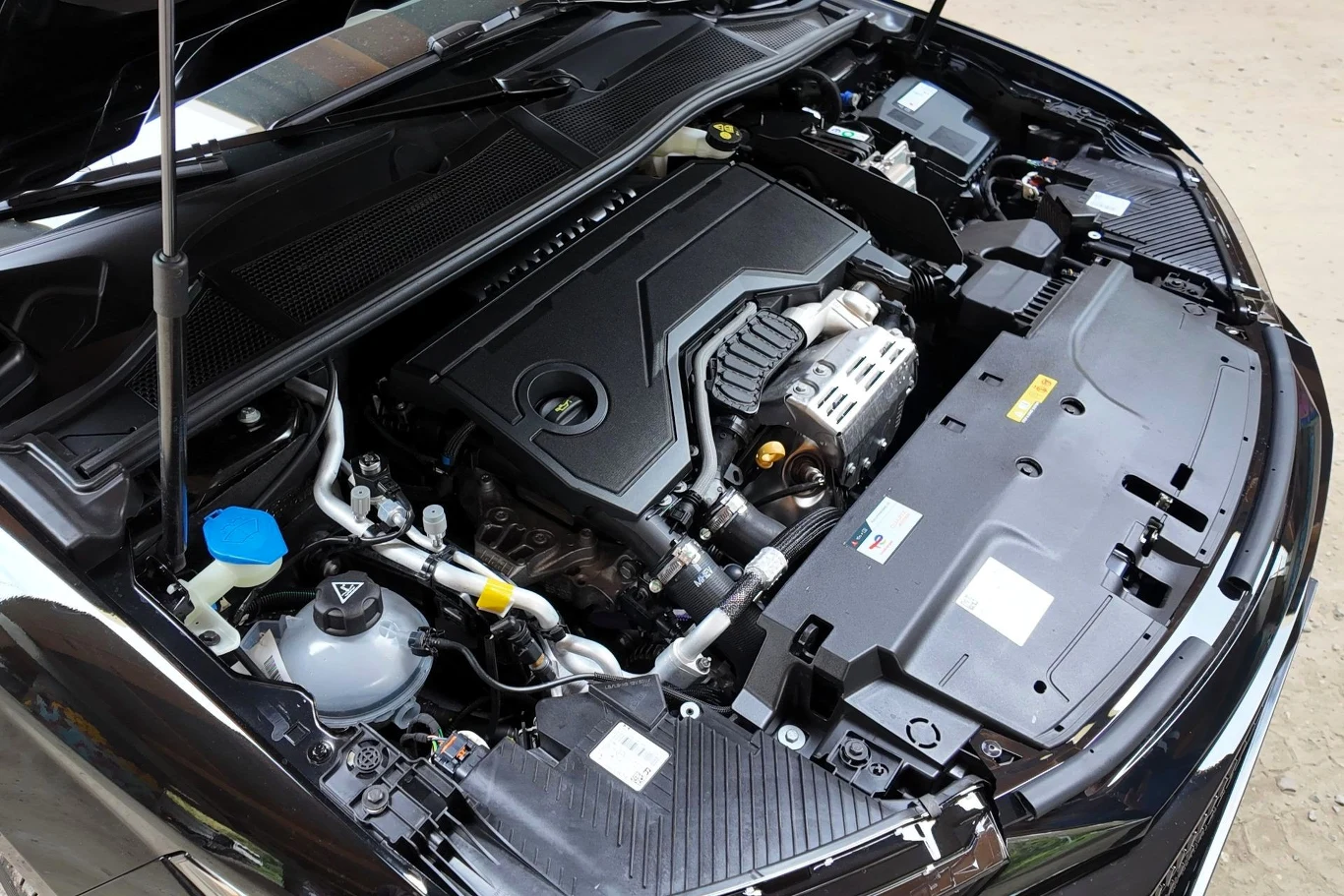 Silnik samochodu osobowego widoczny pod otwartą maską, wyraźnie pokazane elementy mechaniczne, osłony oraz zbiornik płynu do spryskiwaczy.