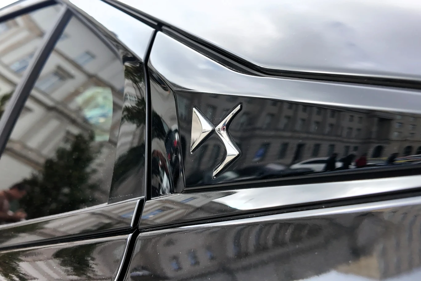 Logo marki DS Automobiles na błyszczącej czarnej karoserii samochodu, w tle odbicie budynku.