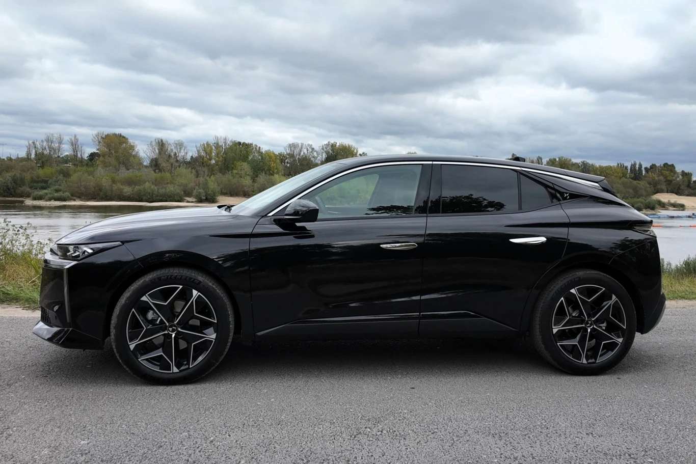 Czarny, nowoczesny SUV zaparkowany na asfalcie z widokiem na rzekę i drzewa w tle, pod pochmurnym niebem.