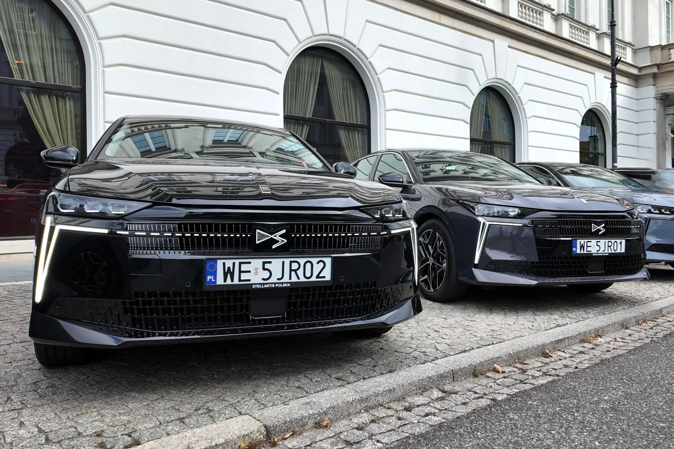 Dwa nowoczesne, czarne samochody elektryczne Xpeng P7 zaparkowane obok siebie na brukowanym chodniku, na tle klasycznego budynku z białą elewacją i łukowymi oknami.