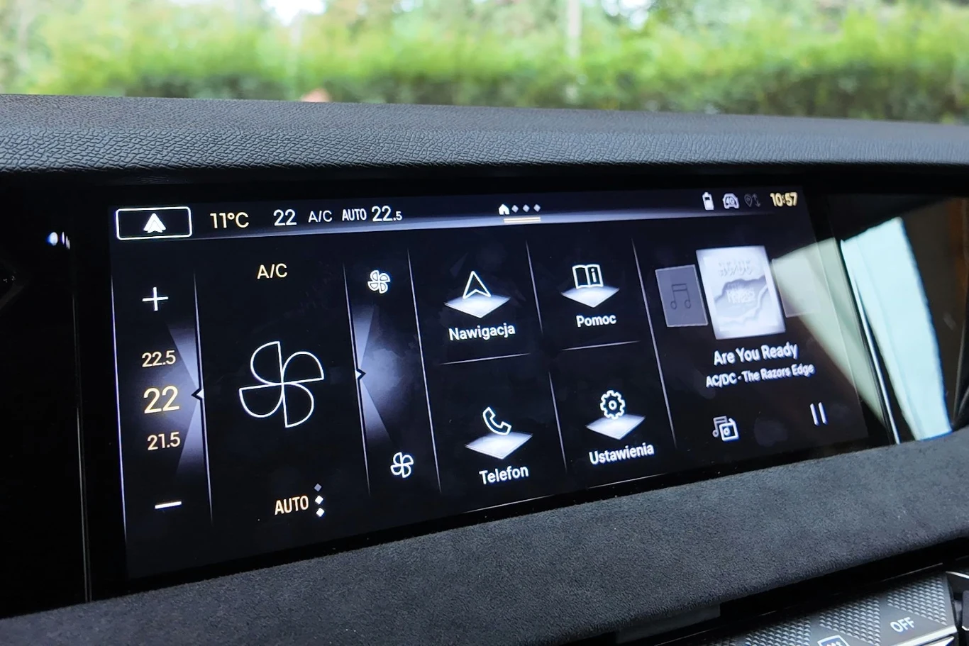 Panel dotykowy samochodu zawierający sterowanie klimatyzacją, nawigację, panel muzyczny z okładką utworu oraz funkcje telefonu i ustawienia, wszystko na jednym szerokim ekranie LCD, z czytelnymi ikonami oraz temperaturami po bokach.