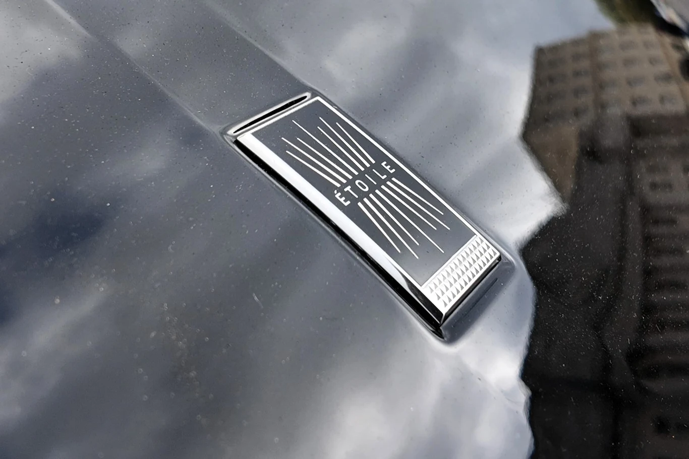 Metalowa plakietka z napisem 'Etoile' zamontowana na błyszczącej czarnej powierzchni samochodu, odbijającej chmury i fragmenty otoczenia.