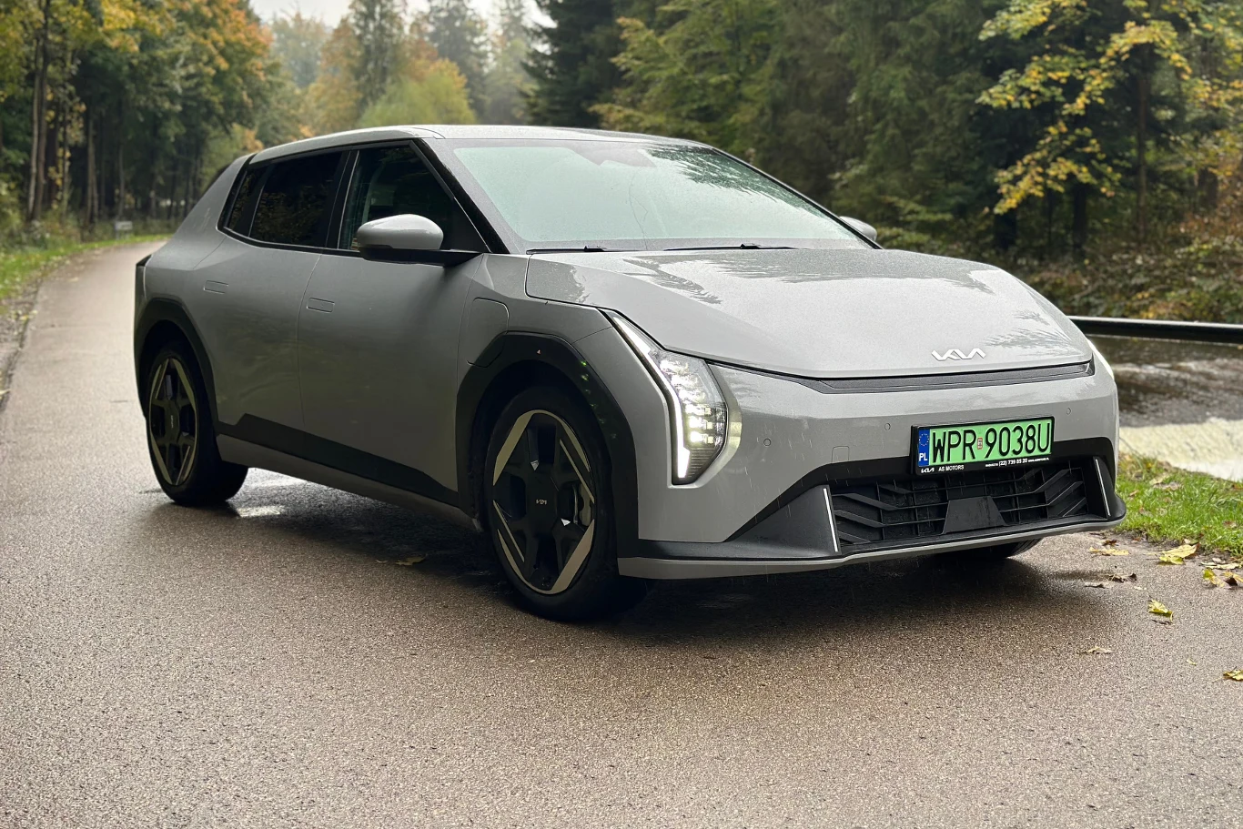 Kia EV4 - elektryczny model skrojony na miarę statystycznego Europejczyka Srebrny samochód elektryczny marki Kia stoi na wilgotnej asfaltowej drodze w otoczeniu zielonego lasu z widocznymi jesiennymi liśćmi na poboczu.