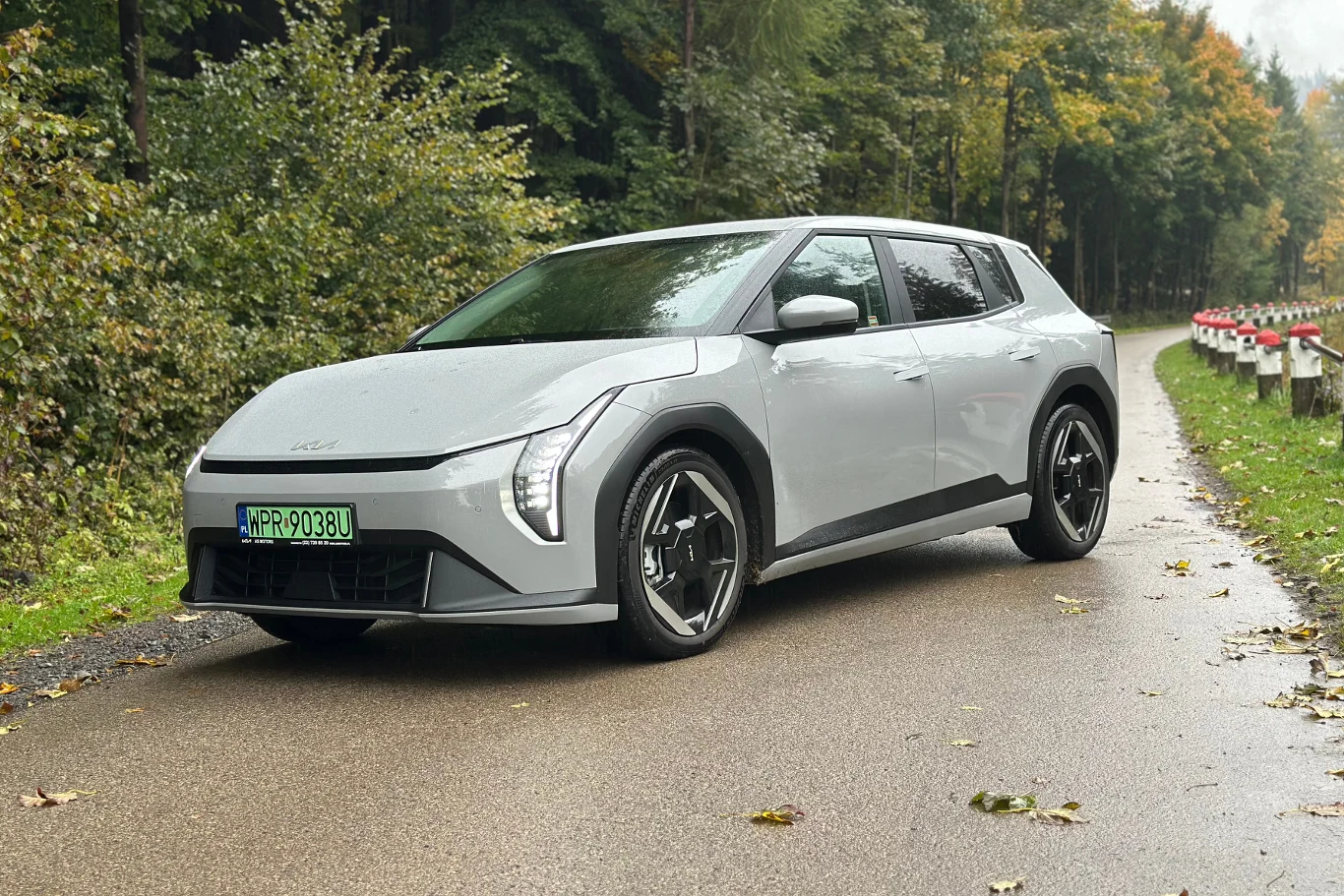 Kia EV4 w wersji hatchback Jasnoszary samochód elektryczny nowoczesnego designu stoi na wilgotnej, asfaltowej drodze otoczonej zielonym lasem. Auto posiada charakterystyczne reflektory LED i czarne felgi. W tle widoczna barierka ochronna i drzewa pokryte jesiennymi liśćmi.