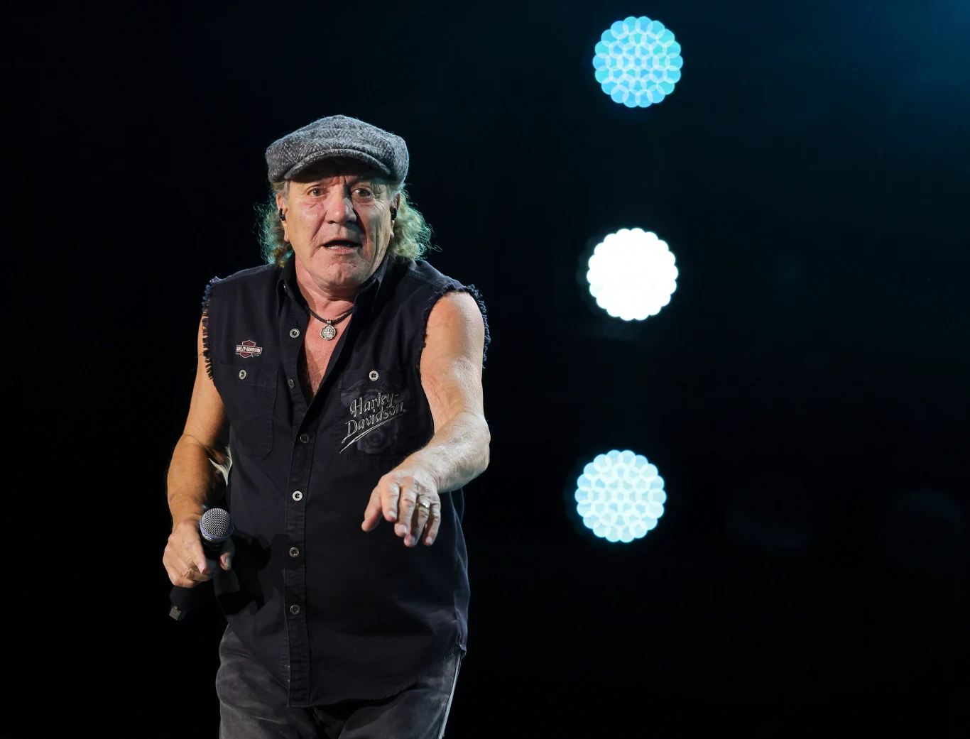 Brian Johnson został wokalistą AC/DC po śmierci Bona Scotta Starszy mężczyzna w czapce z daszkiem i kamizelce bez rękawów trzyma mikrofon, stoi na scenie na tle jasnych reflektorów scenicznych.