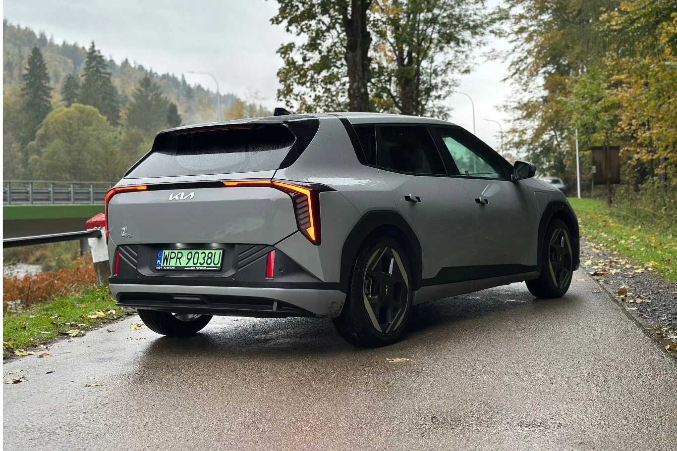 Bagażnik EV4 hatchback ma pojemność 435 litrów Nowoczesny szary samochód marki Kia o futurystycznym designie stoi na mokrej asfaltowej drodze otoczonej drzewami, z widocznymi pomarańczowymi światłami i zielonymi tablicami rejestracyjnymi.