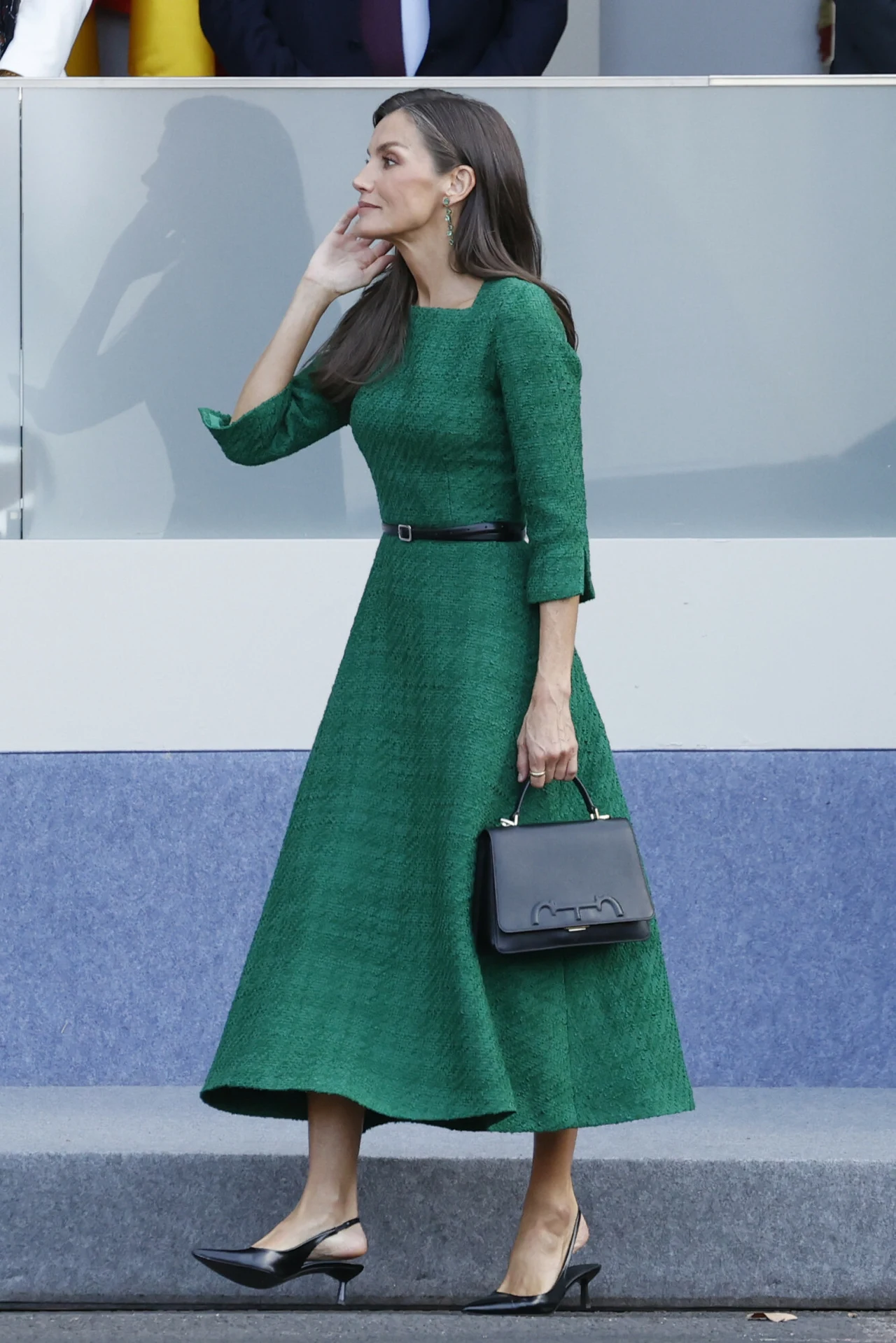 Królowa Letycja podczas obchodów Narodowego Święta Hiszpanii Elegancka kobieta w zielonej sukience do połowy łydki z czarnym paskiem, trzymająca czarną torebkę i nosząca czarne buty na obcasie, idzie chodnikiem obok przeszklonej balustrady.
