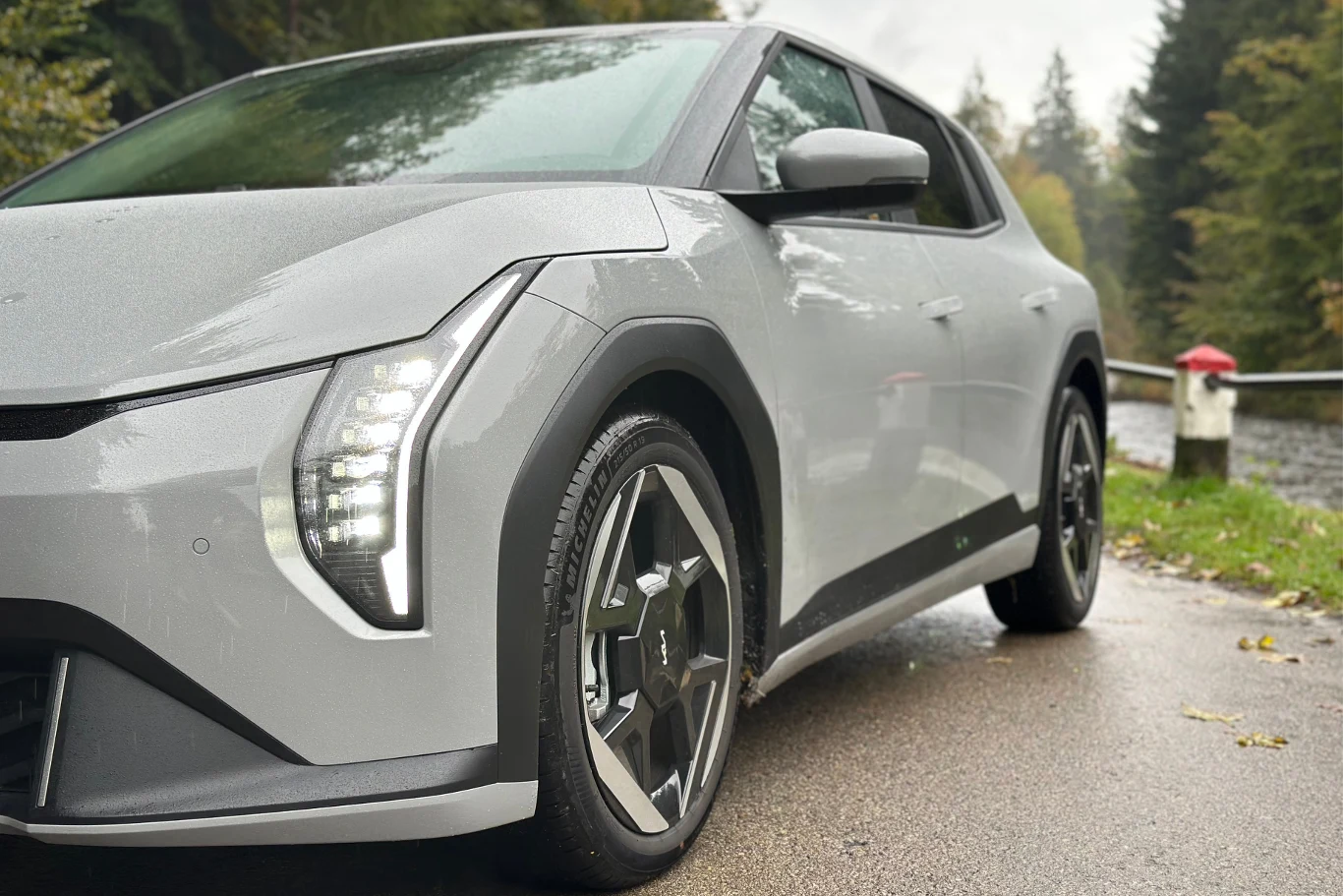 Klamki zostały ukryte - wielu twierdzi, że nie jest to najbardziej użyteczne rozwiązanie, jednakże w tym przypadku chodziło o aerodynamikę Nowoczesny szary samochód elektryczny zaparkowany na wilgotnej drodze w otoczeniu leśnego krajobrazu, z widocznym nowoczesnym oświetleniem LED i sportowymi felgami