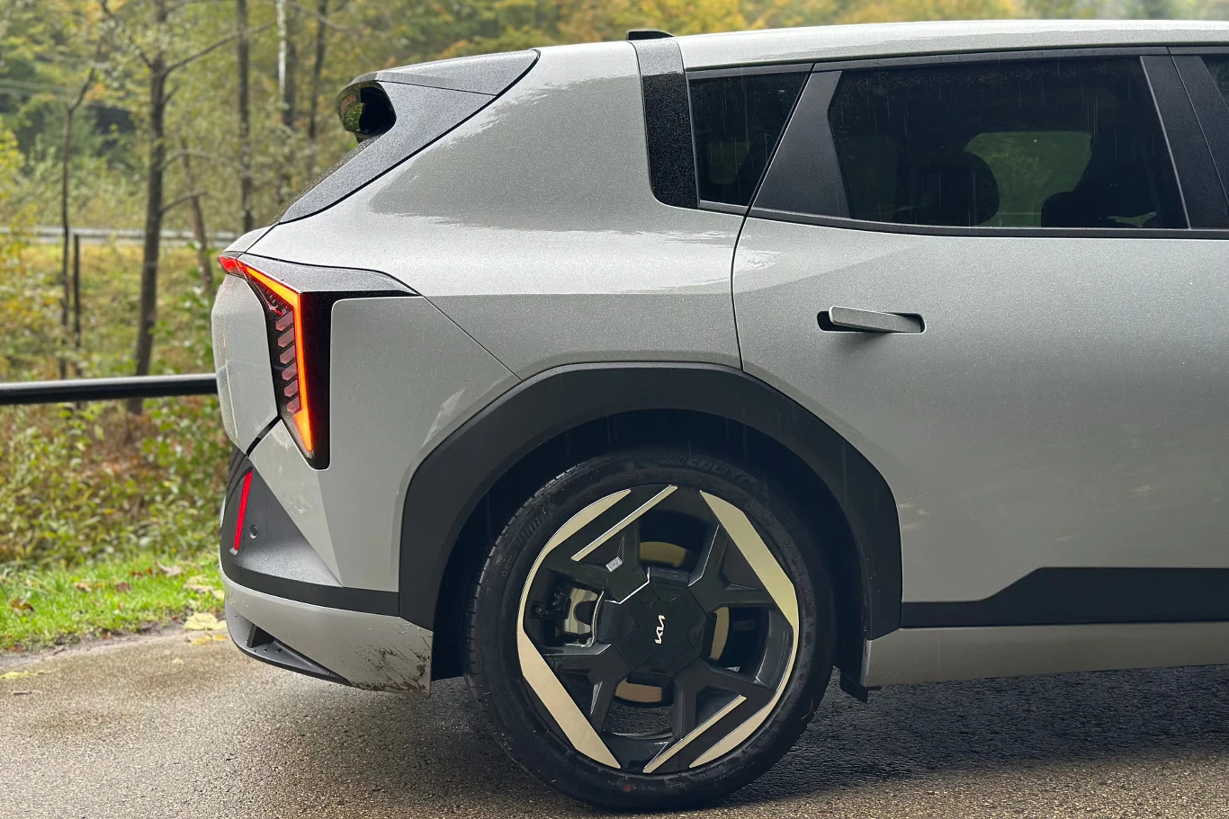 Kia EV4 w wersji hatchback Tylna część srebrnego samochodu elektrycznego z nowoczesnymi, dynamicznie zaprojektowanymi lampami LED i felgami. Auto stoi na wilgotnej nawierzchni przy zielonym tle leśnym.